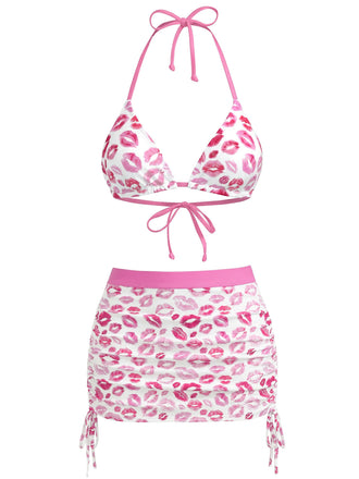 Ein dreiteiliges Bademodenset bestehend aus einem weißen Triangel-Bikini-Oberteil, passendem Bikinihöschen mit Bindebändern an den Seiten und einem gerafften Baderock, alle mit einem pinken Lippenstiftkuss-Print.