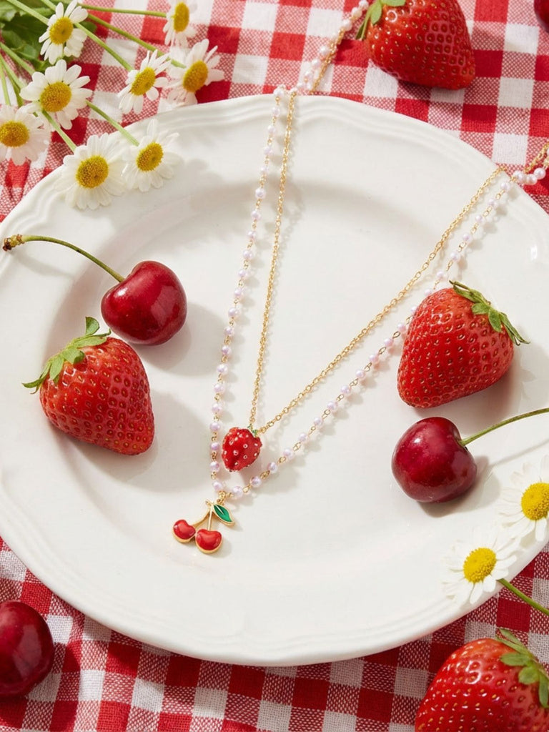 Halskette mit Erdbeer- und Kirschmotiven auf einem weißen Teller mit Erdbeeren und Kirschen auf einer rot-weiß karierten Tischdecke.