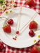 Halskette mit Erdbeer- und Kirschmotiven auf einem weißen Teller mit Erdbeeren und Kirschen auf einer rot-weiß karierten Tischdecke.