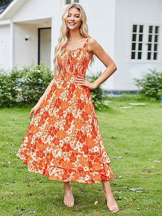 Eine Frau in einem orangefarbenen Blumenkleid steht auf einer Rasenfläche; im Hintergrund ist ein Haus zu sehen.