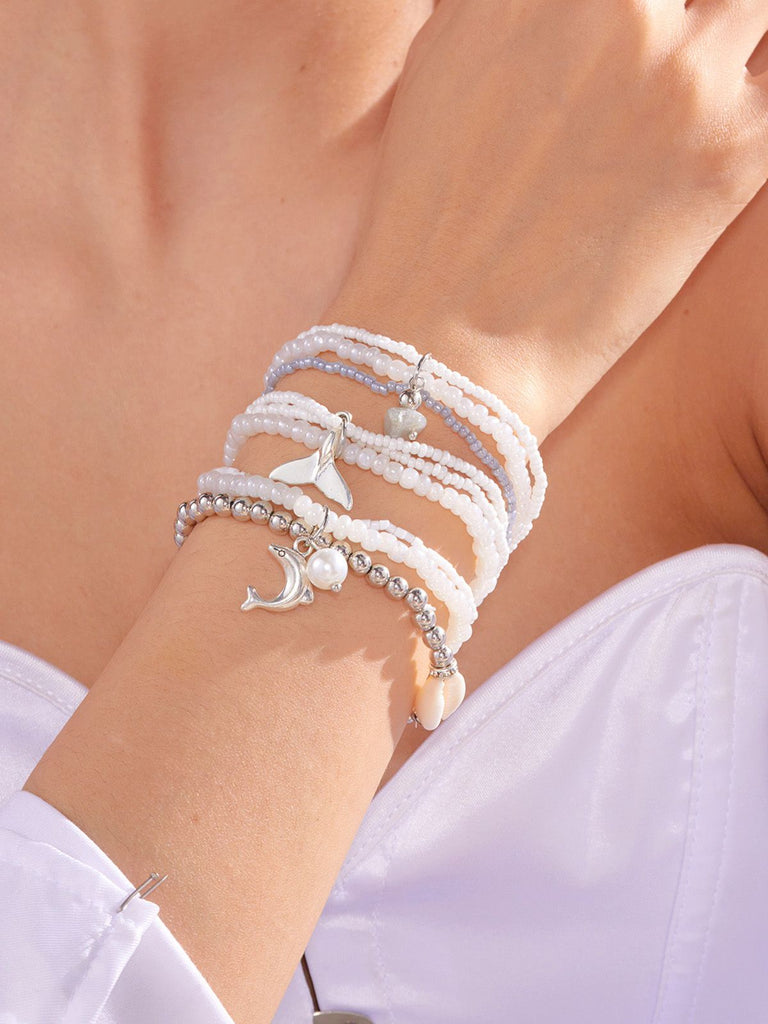 Nahaufnahme eines Handgelenks mit mehreren weißen Perlenarmbändern mit silbernen Ankeranhängern.